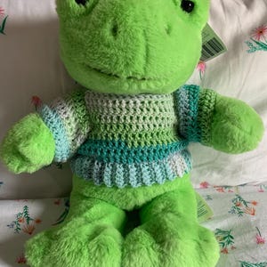 Puede incluir: Un peluche de rana verde con un suéter de ganchillo en tonos verdes, azules y blancos. La rana tiene ojos negros grandes y un cuerpo suave y texturizado. El juguete está sobre una superficie con estampado floral.