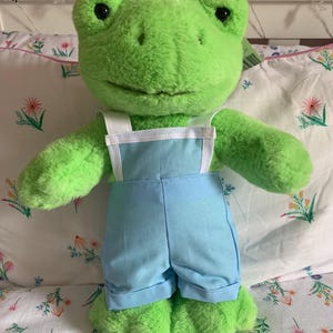 Puede incluir: Un peluche de rana verde con grandes ojos negros y una sonrisa. La rana lleva un peto azul claro con tirantes blancos. El juguete está colocado sobre una almohada con estampado floral.