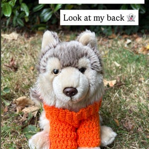May include: A plush wolf toy wearing a bright orange crocheted sweater. The toy has grey and white fur, dark eyes, and a brown nose. Text overlays on the image state: Webkinz not included! Look at my back.