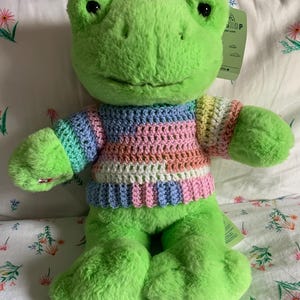 Puede incluir: Un peluche verde con forma de rana que lleva un suéter de ganchillo a rayas multicolores. El suéter tiene rayas rosas, azules y amarillas. La rana tiene grandes ojos negros y está sentada.