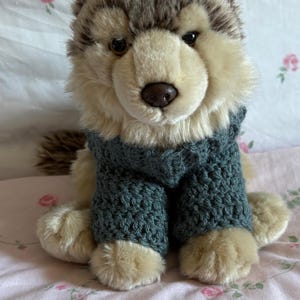May include: A plush wolf toy with tan and brown fur wearing a dark teal crocheted sweater. The toy has dark brown eyes and a dark nose. A white rectangular label in the upper right corner contains the text: Webkinz signature not included.
