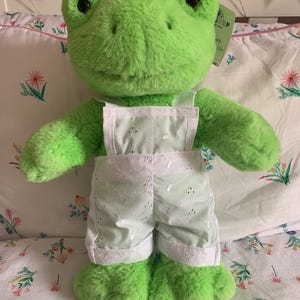 Puede incluir: Un peluche verde con forma de rana que lleva un peto blanco con un diseño floral. La rana tiene grandes ojos negros y está colocada sobre una colcha con estampado floral. Tiene una etiqueta.