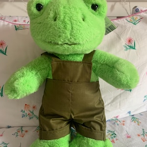 Puede incluir: Un peluche verde con forma de rana que lleva un mono marrón. La rana tiene ojos negros y una expresión amigable. El juguete de peluche está diseñado para niños.