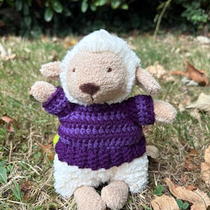 May include: A plush lamb toy with cream-colored fur and a dark brown nose, wearing a hand-crocheted short-sleeved sweater in a deep purple yarn. The stuffed animal has floppy ears, a soft textured body, and is positioned in a seated posture.