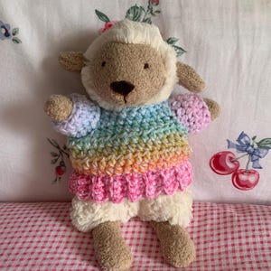 May include: A plush lamb stuffed animal with tan fur and a cream-colored face. The lamb is wearing a crocheted sweater featuring horizontal stripes of pastel rainbow colors, including blue, yellow, orange, and pink, with a ribbed pink hem.
