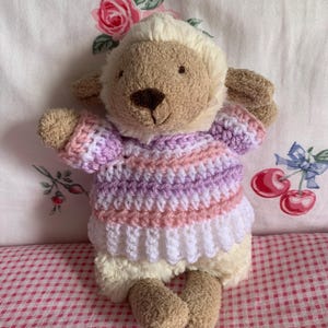 May include: A plush lamb stuffed animal with cream and tan fur, wearing a hand-crocheted sweater featuring horizontal stripes in shades of light purple, soft pink, and white. The toy has a dark brown nose and stitched facial features.