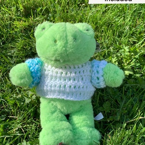 Puede incluir: Rana de peluche verde con un suéter tejido de color blanco y azul. La rana está sentada en un lecho de hierba verde.