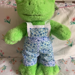 Puede incluir: Una rana de peluche verde con un peto azul con estampado floral y tirantes blancos. La rana tiene grandes ojos negros y está de pie sobre una superficie con estampado floral. Una etiqueta está adherida a la rana.