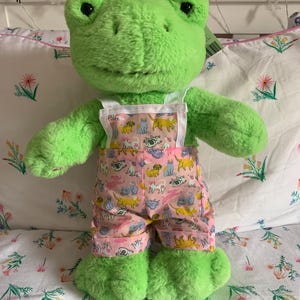 Puede incluir: Una rana de peluche verde con un mono rosa estampado con perros y gatos de dibujos animados. La rana tiene grandes ojos negros y está sobre una colcha floral. El mono tiene tirantes blancos.