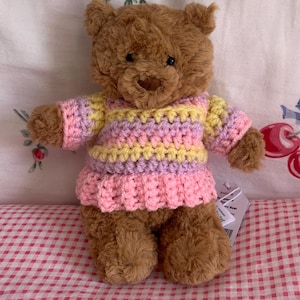 Op de afbeelding: Een bruine pluche teddybeer met een handgehaakte trui met horizontale strepen in pastelgeel, lavendel en roze. De beer heeft donkere ogen en een bruine neus, en een klein wit productlabel is aan de zijkant bij de arm bevestigd.