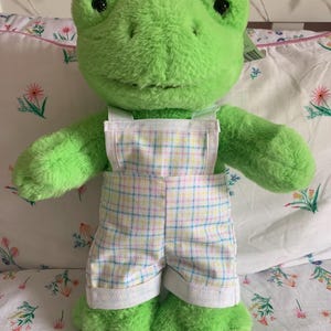 Puede incluir: Un juguete de peluche verde con forma de rana, con ojos negros y una sonrisa. La rana lleva un peto a cuadros en colores pastel. El juguete está sobre una colcha con estampado floral.