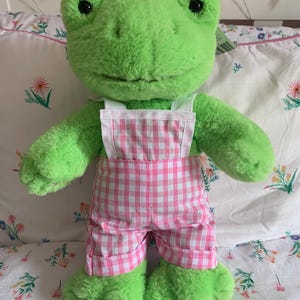 Puede incluir: Un peluche verde con forma de rana que lleva un peto rosa y blanco a cuadros. La rana tiene grandes ojos negros y una expresión amistosa. El peluche está sobre una colcha con estampado floral.