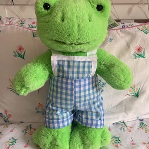 Puede incluir: Un juguete de peluche verde con forma de rana que lleva un peto a cuadros azules y blancos. La rana tiene ojos grandes y redondos y una expresión amigable. El juguete está hecho de material suave y difuso.