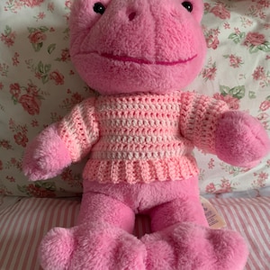 May include: A pink plush frog wearing a pink and white crocheted sweater. The frog is sitting on a striped pink and white pillow.