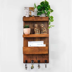 May include: A rustic wooden wall organizer with three shelves and a key rack. The top shelf holds a small plant and decorative items. The middle shelf displays two figurines and a small potted plant. The bottom shelf has a mail slot and a key rack with several keys.