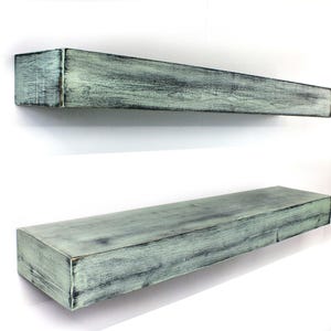 May include: Two distressed, rectangular floating shelves. The shelves are painted in a muted teal color with a weathered, textured finish. The shelves are mounted on a white wall, showcasing their rustic charm and design.