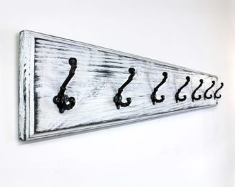 May include: A distressed white wooden coat rack with six black metal hooks. The rack has a rectangular shape and is mounted on a white wall. The wood has a weathered appearance, with visible grain and a distressed white finish.
