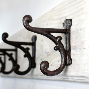 May include: Close-up of a decorative, dark brown metal coat hook with a curved design. The hook is mounted on a distressed, white-painted wooden board. The hook has a scroll-like design and is attached with screws.
