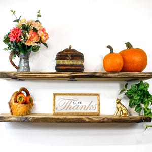 May include: Two rustic wooden shelves with various decorative items. The top shelf holds a metal pitcher with flowers, a wooden box, and two pumpkins. The bottom shelf displays a basket of fruit, a framed sign that says "Give Thanks", and a golden cat statue.