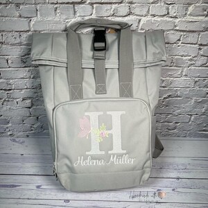 Könnte beinhalten: Grauer Rucksack mit einer Vordertasche, bestickt mit dem Buchstaben "H" mit Blumen- und Schmetterlingsdesign und dem Namen "Helena Müller" darunter. Der Rucksack hat einen oberen Griff und verstellbare Träger.