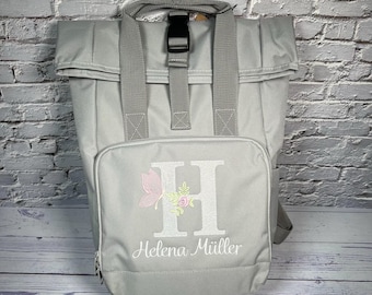 Könnte beinhalten: Grauer Rucksack mit einer Vordertasche, bestickt mit dem Buchstaben "H" mit Blumen- und Schmetterlingsdesign und dem Namen "Helena Müller" darunter. Der Rucksack hat einen oberen Griff und verstellbare Träger.