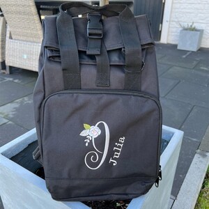 Könnte beinhalten: Schwarzer Rucksack mit Reißverschluss und verstellbaren Trägern. Der Rucksack hat ein florales Design und den Namen "Julia" auf der Vorderseite gestickt.