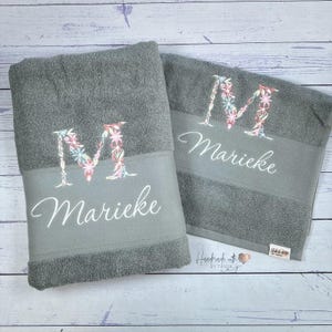 Könnte beinhalten: Zwei graue Badetücher mit einem großen, mit bunten Blumenmustern bestickten Buchstaben M und dem Namen Marieke in weißer Schreibschrift. Jedes Handtuch hat ein glattes Band in der Mitte und ein kleines Stoffetikett an der Ecke.