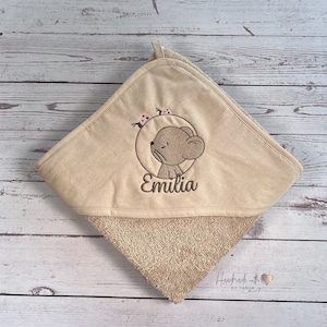 Puede incluir: Toalla con capucha beige con un oso marr&oacute;n bordado y el nombre "Emilia" en ella. La toalla tiene un respaldo de tejido de felpa marr&oacute;n.