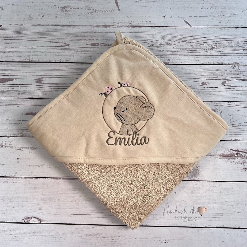 Puede incluir: Toalla con capucha beige con un oso marr&oacute;n bordado y el nombre "Emilia" en ella. La toalla tiene un respaldo de tejido de felpa marr&oacute;n.