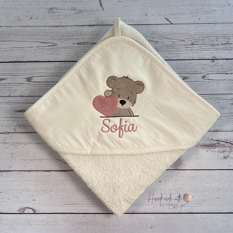Puede incluir: Una toalla con capucha de color crema con un oso de peluche marr&oacute;n que sostiene un coraz&oacute;n rosa. La toalla est&aacute; bordada con el nombre "Sofia".