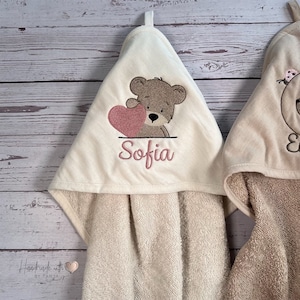 Puede incluir: Dos toallas con capucha con osos de peluche bordados. Una toalla presenta un oso de peluche marr&oacute;n que sostiene un coraz&oacute;n rosa y el nombre "Sofia" bordado debajo. La otra toalla presenta un oso de peluche marr&oacute;n con una flor rosa en la cabeza.