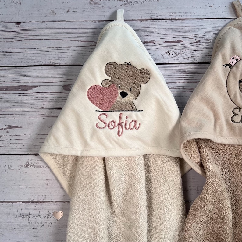 Puede incluir: Dos toallas con capucha con osos de peluche bordados. Una toalla presenta un oso de peluche marr&oacute;n que sostiene un coraz&oacute;n rosa y el nombre "Sofia" bordado debajo. La otra toalla presenta un oso de peluche marr&oacute;n con una flor rosa en la cabeza.