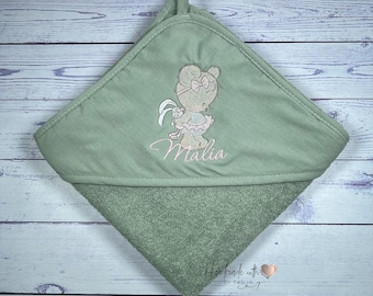 May include: Sage green hooded baby towel with an embroidered bear design holding a bunny and the name "Malia" in cursive. The towel is folded on a light wood-grain surface. The text "Handmade with love by Tanja" is visible.
