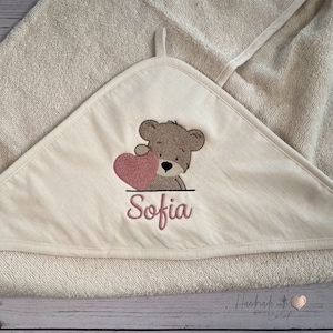 Puede incluir: Una toalla con capucha beige con un oso marr&oacute;n que sostiene un coraz&oacute;n rosa y el nombre "Sofia" bordado. La toalla est&aacute; hecha a mano con un logotipo de coraz&oacute;n.