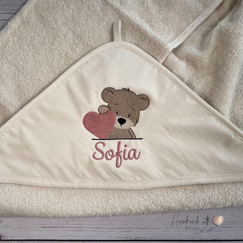 Puede incluir: Una toalla con capucha beige con un oso marr&oacute;n que sostiene un coraz&oacute;n rosa y el nombre "Sofia" bordado. La toalla est&aacute; hecha a mano con un logotipo de coraz&oacute;n.