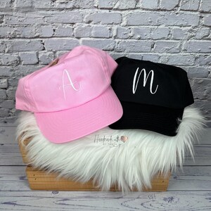 Könnte beinhalten: Zwei Baseballkappen, eine rosa und eine schwarze, liegen auf weißem Kunstfell in einer Holzkiste. Die rosa Kappe hat ein weiß gesticktes "A", die schwarze Kappe ein weiß gesticktes "M". Der Hintergrund ist eine weiße Backsteinmauer.