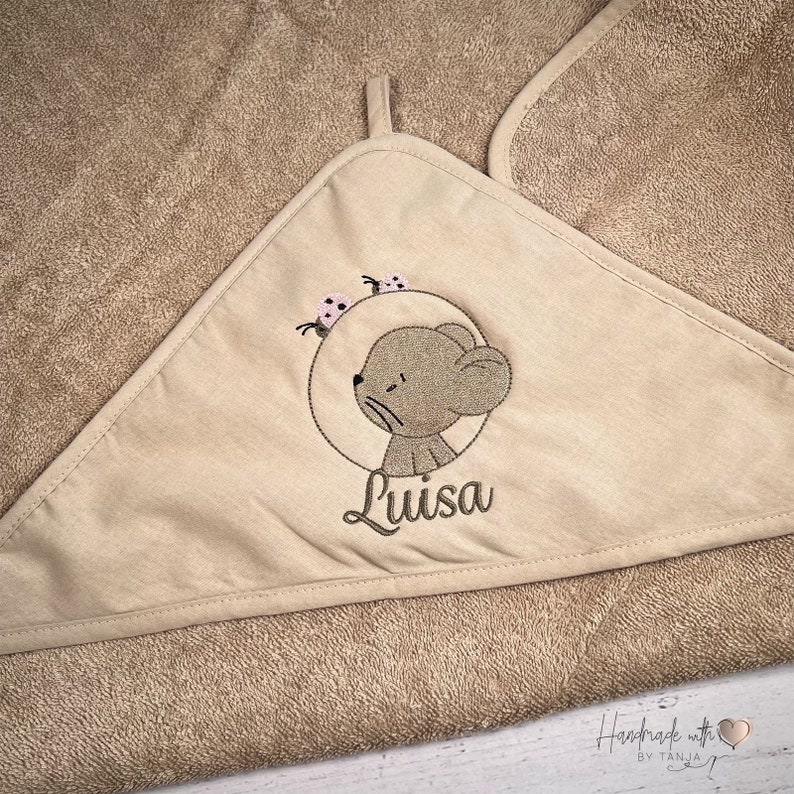 Puede incluir: Una toalla con capucha beige con un rat&oacute;n bordado marr&oacute;n y el nombre "Luisa" en ella. El rat&oacute;n est&aacute; rodeado de tres mariquitas rosas. La toalla tiene el texto "Handmade with love by Tanja" en la esquina inferior derecha.