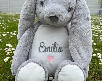 May include: A gray plush bunny with long ears and a white patch on its chest. The patch has the name "Emilia" embroidered on it in gray thread, with a pink heart above the name.