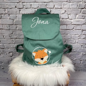 Könnte beinhalten: Grüner Rucksack mit Klappenverschluss, bestickt mit dem Namen "Jona" in weißer Schrift. Auf der Vorderseite befindet sich ein kreisförmiger Aufnäher mit einer orangefarbenen Fuchsdarstellung. Der Rucksack hat einen oberen Griff und verstellbare Träger.