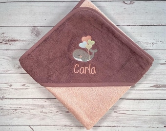 May include: A brown hooded towel with a light pink lining. The towel has an embroidered whale with balloons and the name "Carla" on it.