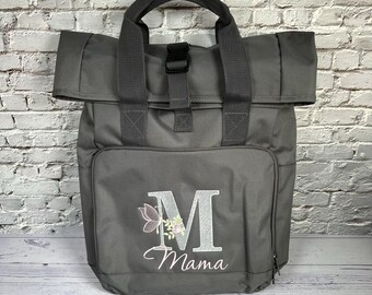 Könnte beinhalten: Grauer Rucksack mit Rollverschluss und Tragegriff. Die Vordertasche hat ein gesticktes Blumendesign mit dem Buchstaben "M" und dem Wort "Mama". Die Tasche hat einen Schnallenverschluss und eine seitliche Reißverschlusstasche.