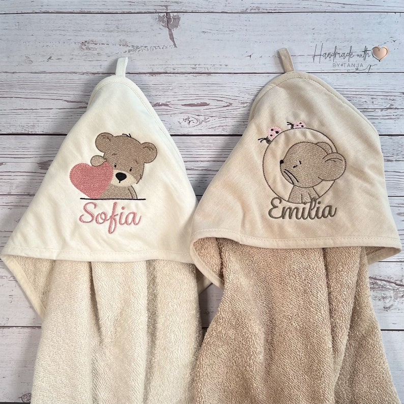 Puede incluir: Dos toallas con capucha con dise&ntilde;os bordados. Una toalla presenta un oso marr&oacute;n sosteniendo un coraz&oacute;n rosa con el nombre "Sofia" bordado debajo. La otra toalla presenta un rat&oacute;n marr&oacute;n con acentos de mariposas rosas y el nombre "Emilia" bordado debajo.
