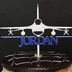 Puede incluir: Topper de pastel de avión blanco con letras azules que dicen "JORDAN". El avión está en vuelo y tiene un fondo blanco. El topper de pastel está encima de un pastel de chocolate.