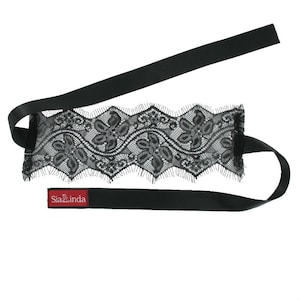 Könnte beinhalten: Schwarze Augenmaske aus Spitze mit floralem Design und schwarzem Satinband. Die Maske hat einen gewellten Rand und ein kleines rotes Etikett mit dem Text "Sia Zinda".