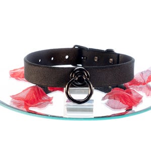 Könnte beinhalten: Schwarzes Choker-Halsband mit großem Metallring. Das Halsband besteht aus einem dunklen, strukturierten Material und verfügt über einen Schnallenverschluss. Der Ring ist ein prominentes Designelement. Das Halsband wird auf einer reflektierenden Oberfläche mit roten Blütenblättern präsentiert.