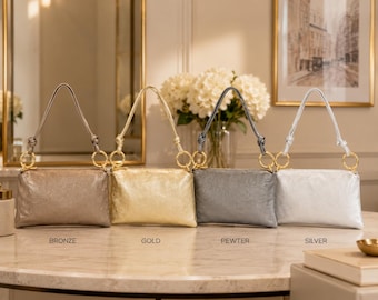May include: Four rectangular metallic handbags arranged in a row, labeled bronze, gold, pewter, and silver. Each bag features a textured finish, a single knotted handle, and gold-toned circular hardware rings connecting the strap to the bag body.
