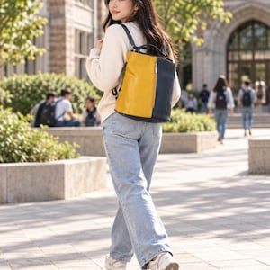 Pode incluir: Uma mochila de dois tons com um design vertical de blocos de cor, apresentando um painel frontal amarelo mostarda e um painel lateral azul-marinho escuro. A bolsa inclui uma alça superior, alças de ombro ajustáveis e um bolso vertical com zíper.