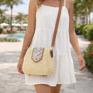 Może przedstawiać: Tkana słomiana torba na ramię z brązowym skórzanym paskiem i klapką ozdobioną kolorowymi kwiatowymi zdobieniami. Torba ma prostokątny kształt i jest noszona na ramieniu.