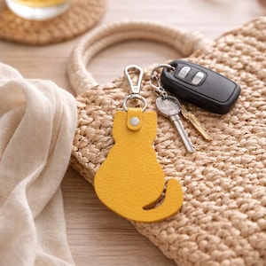 Puede incluir: Un llavero de cuero amarillo en forma de gato con un cierre plateado, junto a un juego de llaves y un mando a distancia de coche. Los artículos descansan sobre un bolso de paja tejida. Gafas de sol y bebida en el fondo.