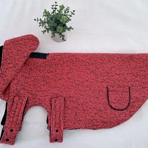 May include: A red and black dog coat with a hood and a pocket. The coat is made of a soft, fleecy material.